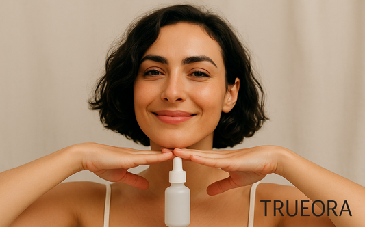 TRUEORA clean beauty model with short wavy hair smiling and holding a white label-free serum bottle between hands under chin