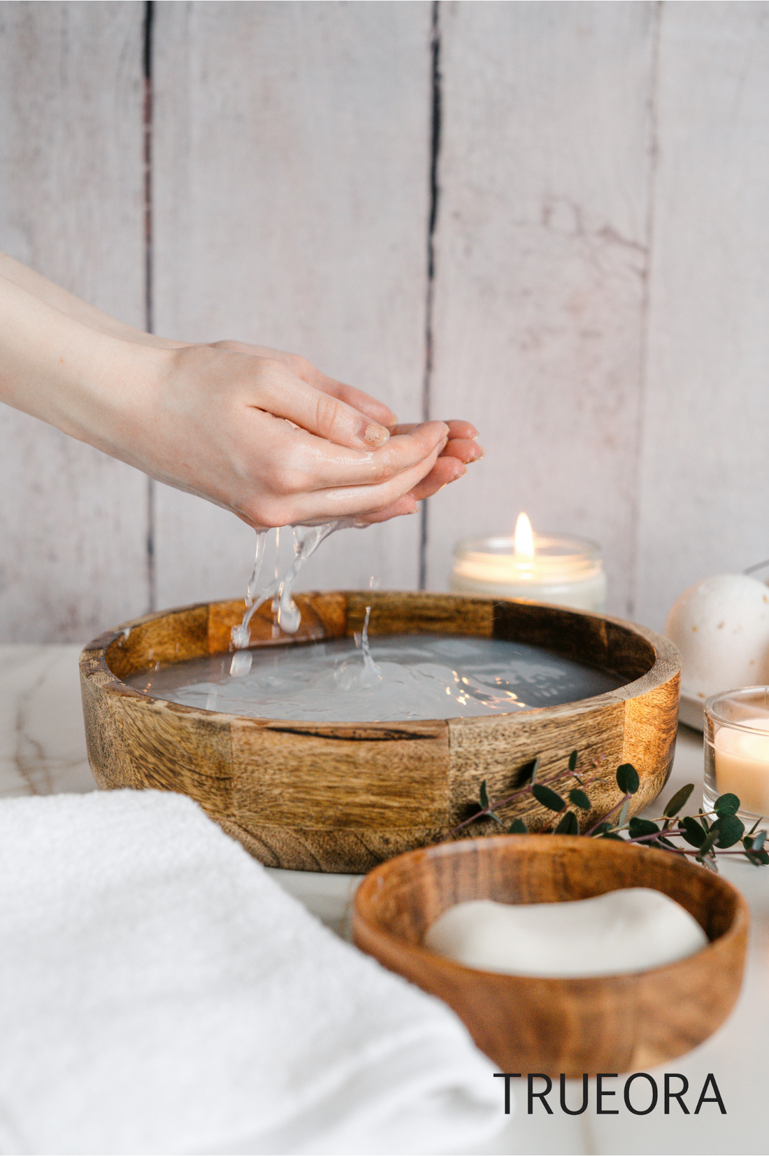 Hands gently cupping water from a wooden bowl in a calm, natural skincare setting with candles, a white towel, and eucalyptus leaves — TRUEORA clean beauty ritual.